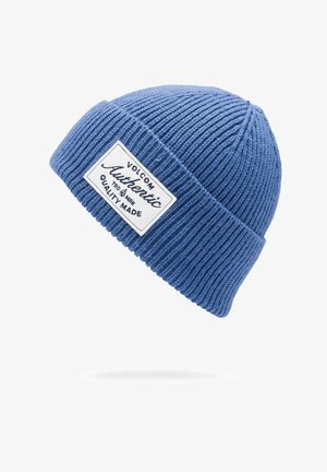 Blauwe geribbelde gebreide beanie met een omgevouwen rand en een rechthoekig wit label met zwarte tekst, met de merknaam en kwaliteitsdetails.