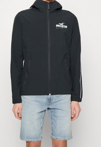 Černá zipová bunda s kapucí, která má bílé logo, dvě zipové kapsy a kontrastní bílé pruhy na rukávech. Nosí se s džínovými šortkami.