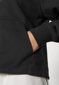Hand resting inside the front pocket of a black sweatshirt with ribbed cuffs, worn over light-colored pants.