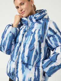 Moteris, vilkinti mėlynai ir balta abstraktaus rašto pūkine striuke su kapišonu, šypsosi ir laiko apykaklę prie veido.