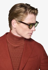 Skildpaddemønstrede solbriller med grønne linser. Modellen bærer en rustfarvet turtleneck og blazer, der fremhæver strukturerede strikkede detaljer og glat stof.
