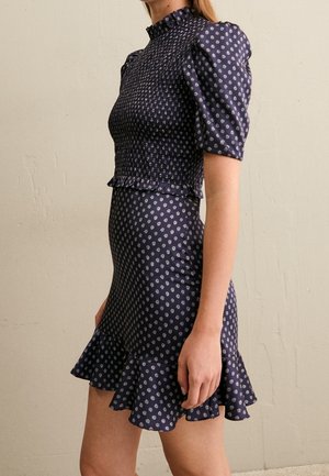 Nainen pukeutuneena tummansiniseen vartalonmyötäiseen mekkoon, jossa on puffy-hihat, smokkaukset yläosassa, pienet valkoiset pilkulliset kuviot ja röyhelöinen helma, seisoo sivuttain.