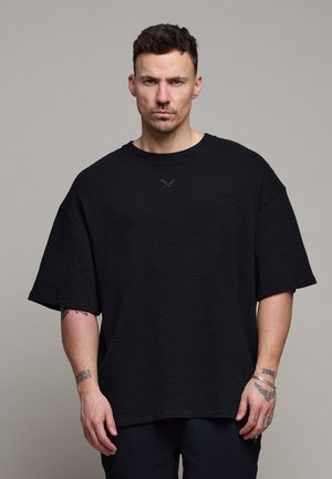 Man met kort donker haar, gekleed in een loszittend zwart textuur kortemouwen shirt en donkere broek, staand tegen een effen grijze achtergrond.