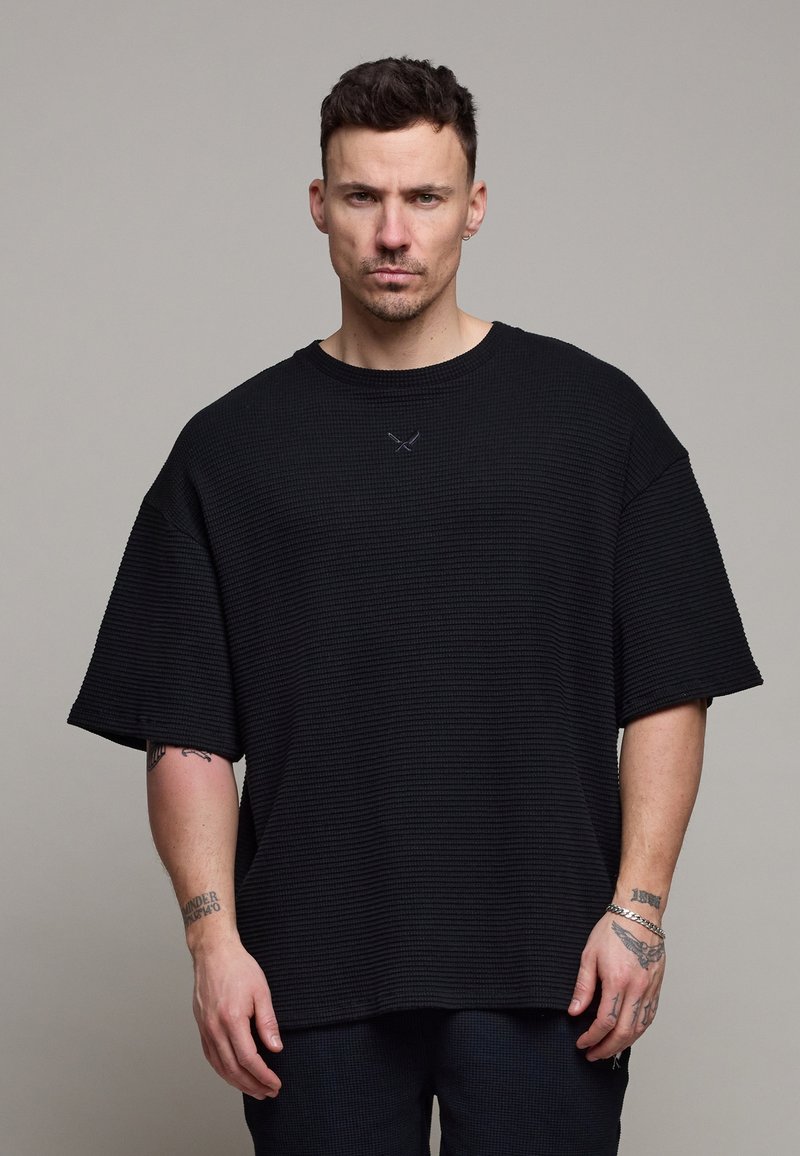 Homme aux cheveux courts et foncés portant une chemise noire texturée à manches courtes et amples, ainsi qu'un pantalon foncé, debout devant un fond gris uni.