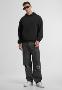 Schwarzer Kapuzenpullover mit einer Fronttasche, kombiniert mit grauen, distressed Jeans und weißen Sneakers. Modell trägt dunkle Sonnenbrillen.