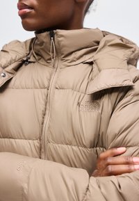 Άτομο που φοράει μπεζ φουσκωτό μπουφάν The North Face με φερμουάρ στον ψηλό γιακά και σταυρωμένα χέρια στο στήθος.