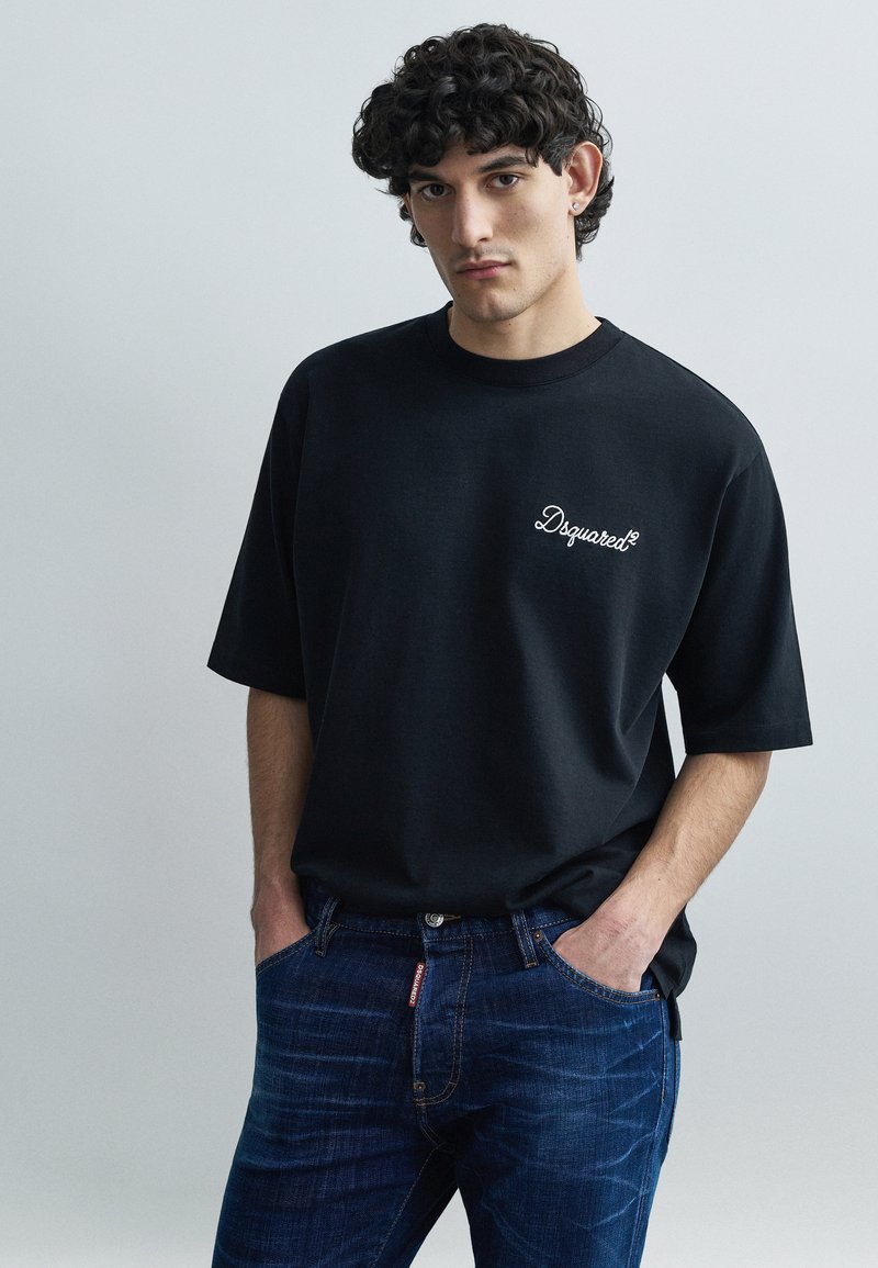 Melnas oversize stila T-krekls ar īsām piedurknēm un izšūtu "Dsquared2" logotipu uz krūtīm, kopā ar tumši ziliem džinsiem.