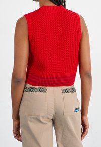 Rode gebreide mouwloze top met een gestructureerd patroon en geribde kraag, gecombineerd met beige broek met patroonaccenten op de zakken.