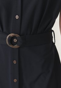 Rochie bleumarin cu talie evidențiată de o curea cu cataramă circulară aurie, detalii cu nasturi pe față și o textură de țesătură netedă.
