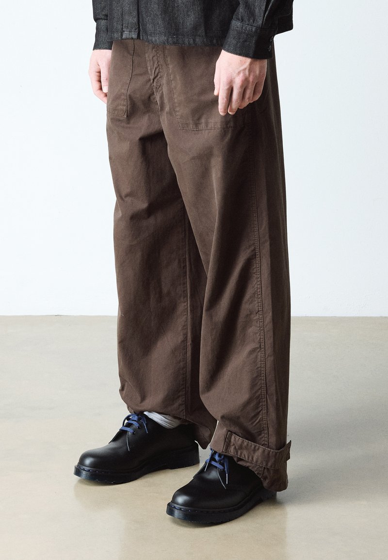 Personne portant un pantalon marron ample avec des poches plaquées et des chaussures noires avec des lacets bleus, debout sur un sol clair.