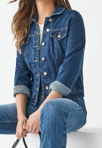 Chaqueta de mezclilla con un diseño ajustado, lavado azul claro y mangas dobladas. Presenta botones de cobre y dos bolsillos en el pecho. Tejido texturizado.