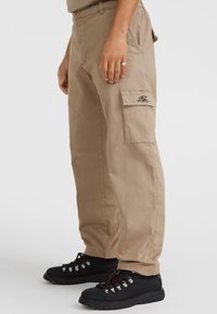 Beige cargo-housut, jotka on valmistettu pehmeästä kankaasta, ja niissä on sivutaskut sekä brodeerattu logo. Yhdistetty mustiin vaelluskenkiin, joissa on punaiset nauhat.