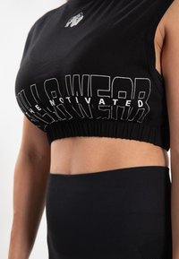 Zwart cropped sporttopje met witte grafische tekst en een logo, voorzien van een geplooemde zoom en een hooggesloten halslijn. Gladde stoftextuur.