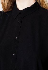 Svart skjorta med knappar och krage, med texturerat tyg och svarta knappar, visas vid axel och övre bröstområde.