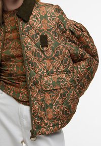 Casaco acolchoado em verde-oliva com padrão floral em laranja. Apresenta um colarinho de tecido de veludo cotelê, fecho com zíper e bolso frontal com botão de pressão.