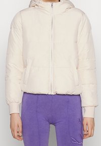 Kvinde iført en lys beige pufferjakke med hætte og lilla bukser med et syet katteansigt design.