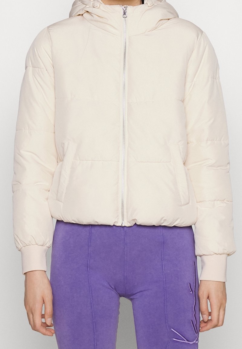 Kvinde iført en lys beige pufferjakke med hætte og lilla bukser med et syet katteansigt design.