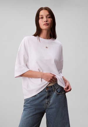 Junge Frau mit langen braunen Haaren, die ein locker sitzendes weißes T-Shirt und blaue Jeans trägt, hält mit einer Hand den Saum ihres Shirts.