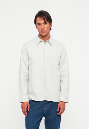 Hombre de pie contra un fondo blanco, vistiendo una camisa blanca de botones de manga larga y jeans azules, de frente con una expresión neutral.