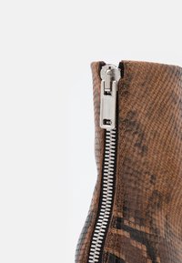 Ruskea käärmeennahkainen nilkkaboots metallic-vetoketjulla ja mustilla yksityiskohdilla, jossa on teksturoitu pinta ja tyylikäs muotoilu.