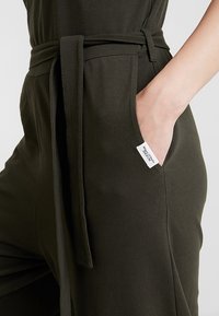Pantalones verde oscuro con bolsillo lateral y cinturón de tela atado a la cintura, mano parcialmente dentro del bolsillo, etiqueta de marca blanca visible.