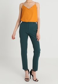 Oranje mouwloze top met een geschulpte halslijn, gecombineerd met groene, op maat gemaakte broek met een slanke pasvorm en zwarte sandalen met hoge hak.