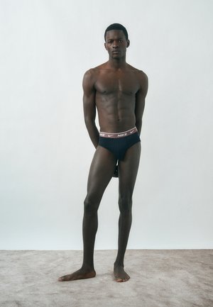 Homme mince et musclé portant un slip noir Nike, debout pieds nus devant un fond clair uni, les mains derrière le dos.