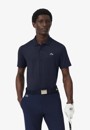 Hombre con camisa tipo polo azul marino y pantalones, sosteniendo un palo de golf con guante blanco, de pie frente a un fondo liso.
