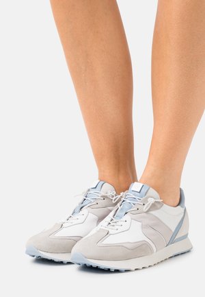 Jambes portant des baskets gris clair et blanches avec des touches de bleu pâle, debout sur un fond clair uni.