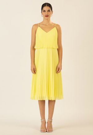 Frau steht, trägt ein ärmelloses, plissiertes gelbes Midikleid mit dünnen Trägern und silbernen Knöchelriemchen-Absätzen vor einfarbigem Hintergrund.