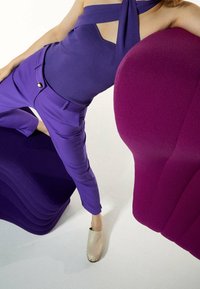 Lila tygtop och byxor, beige skor med metalliska accenter, placerade mot lila och djuprosa skulpturella stolar.