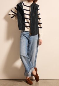 Black sleeveless jacket over a cream striped sweater, paired with light blue wide-leg jeans and brown suede slip-on shoes.