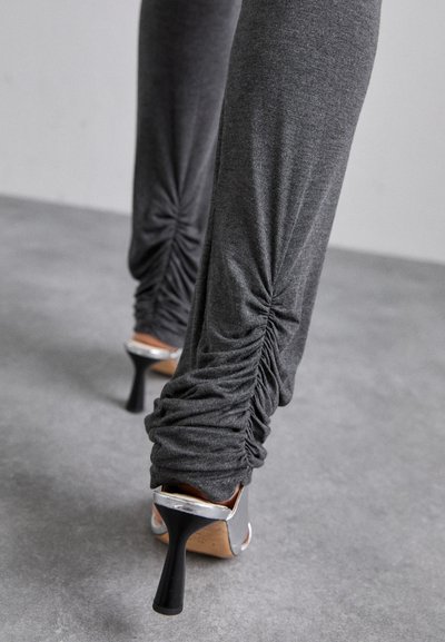 Leggings froncés gris associés à des chaussures à talons argentés. Le tissu a une texture lisse et le design présente des fronces serrées au niveau de la cheville.
