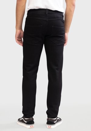 Person wearing black slim-fit jeans and black-and-white sneakers, standing facing away against a plain light background.