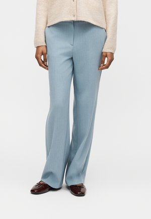 Mujer con pantalones anchos azul claro, cárdigan beige y mocasines de charol marrón oscuro, de pie contra un fondo liso.