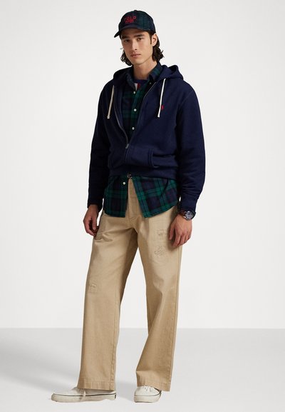 Merihenkinen huppari, jossa on nyörit, vihreä ruudullinen paita alla, beiget leveälahkeiset housut ja valkoiset lenkkarit. Housuissa on minimaalista kulutusta.