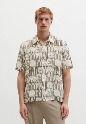 Blonder Mann trägt ein beige-weißes Kurzarmhemd mit abstrakten Blumen- und Geometriemustern, steht vor einem weißen Hintergrund.