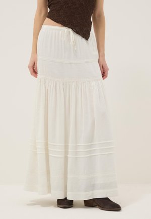 Frau trägt einen langen weißen gestuften Rock mit Spitzendetails, kombiniert mit einem braunen, strukturierten Oberteil und dunkelbraunen Knöchelstiefeln.