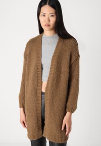 Cárdigan marrón de punto con abertura frontal, que presenta una textura gruesa y acentos de canalé, combinado con una camiseta gris ajustada.