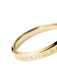 Kultainen bangle-rannekoru, jossa on kaiverrettu teksti "DANIEL WELLINGTON", sileä pinta, pyöreä muoto ja minimalistinen muotoilu.