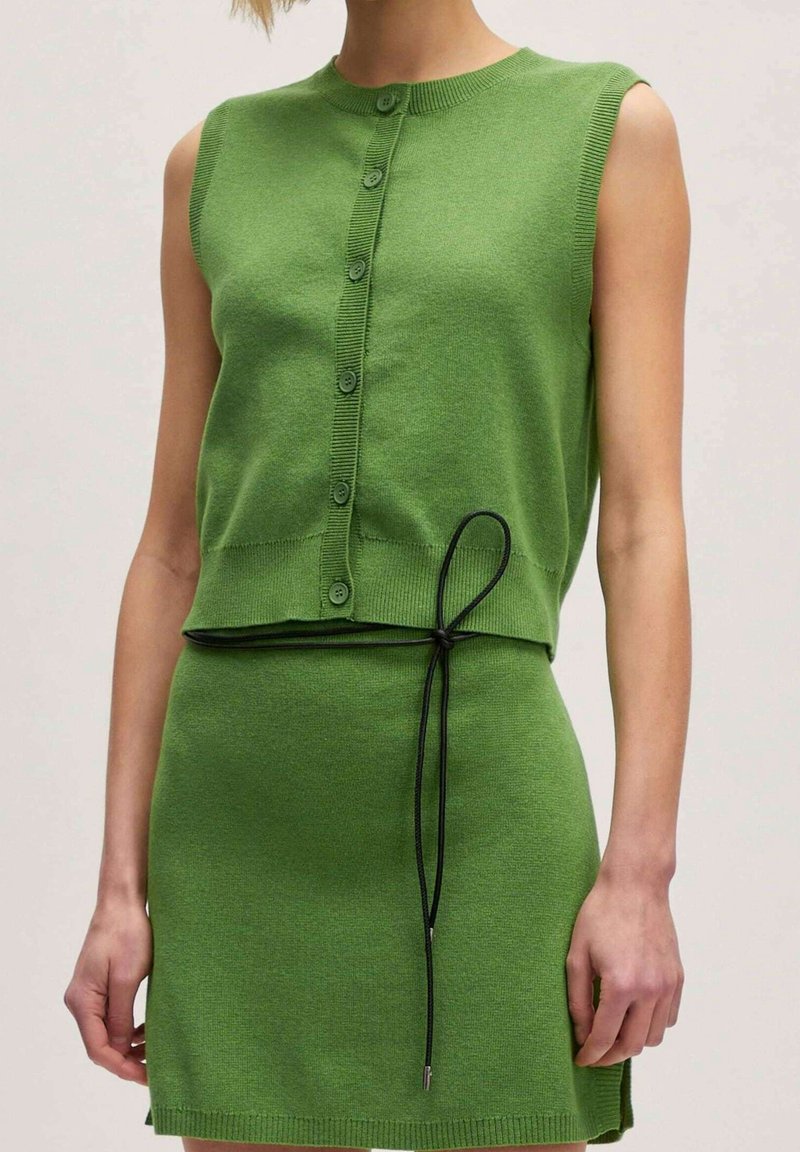 Femme portant un haut vert sans manches boutonné et une jupe verte assortie avec une fine ceinture noire nouée en nœud à la taille.