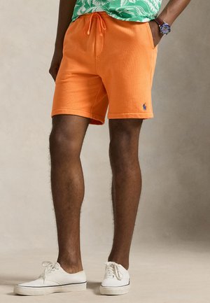 Mężczyzna w pomarańczowych szortach z sznurkiem, zielonej koszuli w wzory, białych trampkach i zegarku na nadgarstku, stojący z jedną ręką w kieszeni.