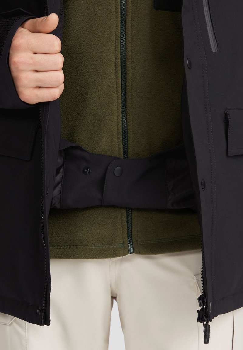 Czarna kurtka zewnętrzna z zamkiem błyskawicznym i zapięciem na napy, odsłaniająca oliwkowo-zieloną wewnętrzną warstwę z polaru. Lekko beżowe spodnie widoczne poniżej.