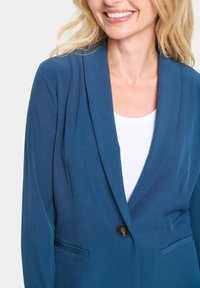 Egy sima anyagból készült, téglalap alakú zöld blézer, egy gombos záródással, kivágott hajtókával és oldalzsebekkel. Fehér felsőre van felvéve.