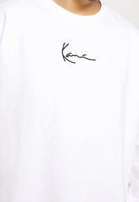 Camisa blanca de manga larga hecha de algodón con cuello redondo. Presenta un logo en escritura negra en el pecho. Textura suave, ajuste informal.
