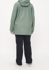 Personne portant une veste à capuche vert clair, un pantalon noir et des baskets blanches, debout de dos sur un fond blanc uni.