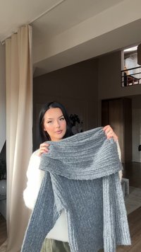 Szary, prążkowany sweter z szerokim, odwróconym dekoltem i długimi rękawami, prezentujący miękką teksturę. Tło w neutralnych odcieniach.