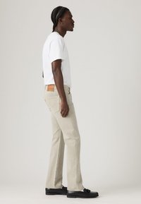 Man with braided hair wearing a white t-shirt, beige straight-leg jeans, white socks, and black loafers standing in profile.