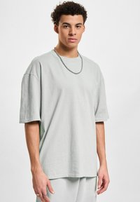 Jasnoszary, oversize bawełniany t-shirt z krótkim rękawem i okrągłym dekoltem, w zestawieniu z srebrnym naszyjnikiem, noszony przez modela.