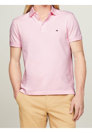 Mężczyzna ubrany w jasnoróżową koszulkę polo z krótkim rękawem i beżowe spodnie, stoi z jedną ręką w kieszeni, widoczny tatuaż na ramieniu.